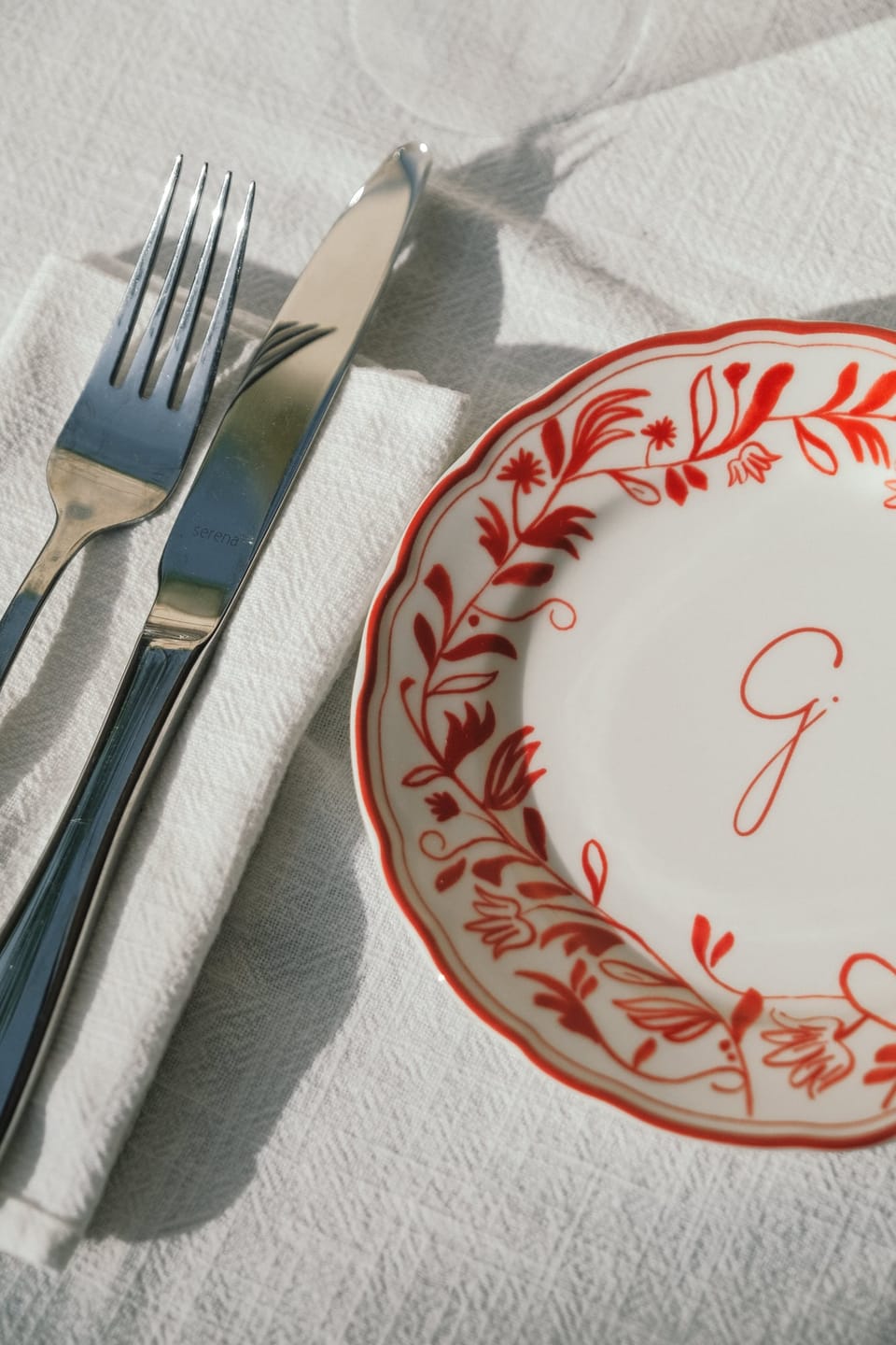 A plate with a red floral rim and the G, with cutlery on white linen