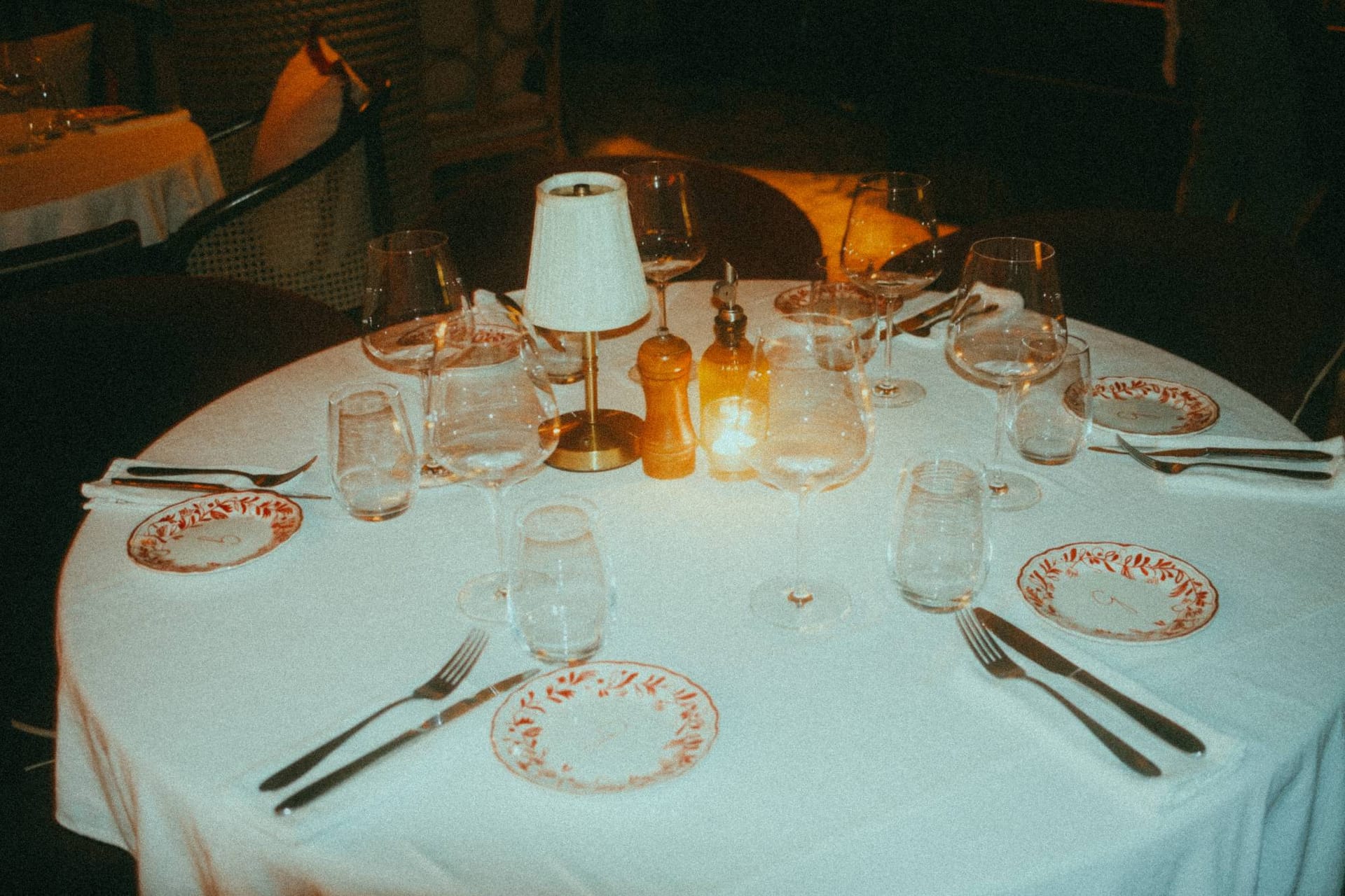 A table set for dinner at night: painted plates, glasses and a small lamp on white linen